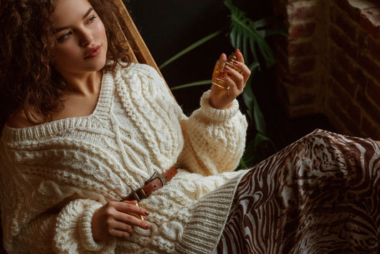 Eine Frau im kuscheligen Pullover lehnt sich im Sessel und hält in der Hand ein Parfüm